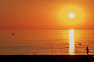 Çocuk bir topla oynuyor ve bir insan plajda gün batımında sörf yapıyor..