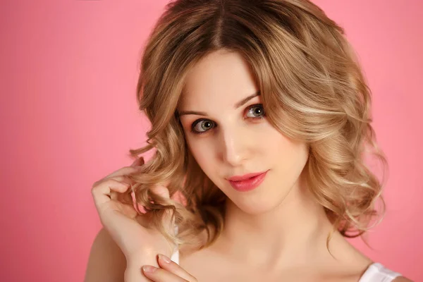 beautiful blonde woman with curly hair in the studio on a pink background, large portrait, face, looking forward, holding hair