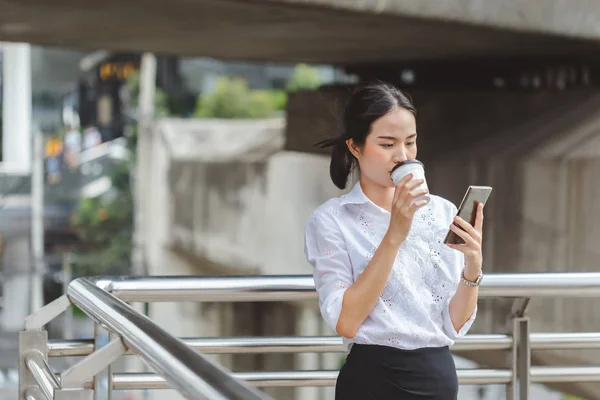 站着的年轻女子白衬衫边喝咖啡边用智能手机干活. — 图库照片