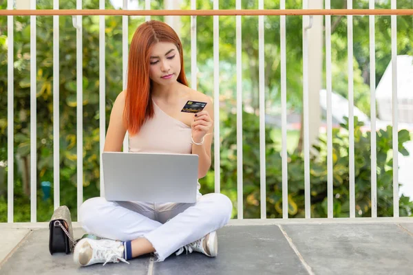 亚洲年轻貌美的女人红头发坐在地板上 手持笔记本电脑 手持信用卡 网上购物购物的理念 — 图库照片