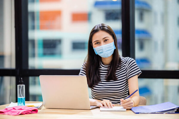 Азиатские женщины носят маски защиты от инфекций, передающихся по воздуху и слюны, во время вспышки вируса Ковид 19 (коронавирус) и работы с ноутбуком в офисе, руки для мытья алкоголя положить на стол.