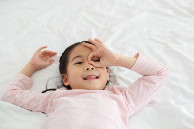 Mutlu küçük kız, beyaz yatağa uzanıp gülümse ve elini gözlerine koy..