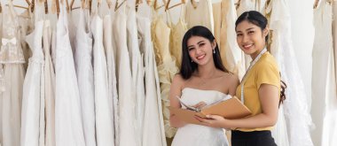 Mutlu Asyalı gelin ve kadın asistan kitap tutuyorlar düğün stüdyosunda gelinlik seçiyorlar, kadın gülümsüyor, düğün konsepti.