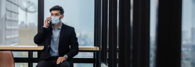 Yakışıklı işadamı maske takıyor. Havadan bulaşan hastalıklara ve tükürük enfeksiyonlarına karşı koruyor. Covid-19 salgını sırasında, ofiste cep telefonuyla konuşuyor..