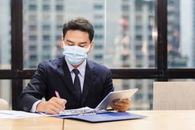 Asyalı genç işadamı dijital tablet kullanarak koruyucu maske takıyor ve ofisteki kağıt işleriyle ilgili veriler yazıyor. Koronavirüs veya covid-19 yayılırken maske takan bir adam..