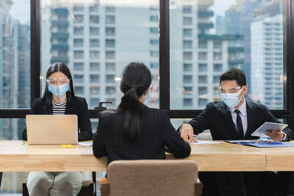 Азиатские бизнесмены надевают маски и защищают лицо от болезней, передающихся по воздуху во время вспышки вируса Ковид-19 или коронавируса, сидя на рабочем социальном расстоянии от работы в офисе.