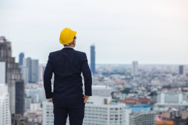 Mimar adam siyah takım elbise giyer ve şehir manzarası ararken arkadan sarı kask takar. Gelecek konsepti aranıyor.