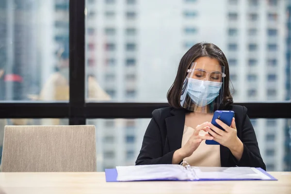 Asian businesswoman wear masks and face shield protect against airborne disease during outbreak of Covid-19 or coronavirus, sitting using smartphone working relaxing moments from work in office.