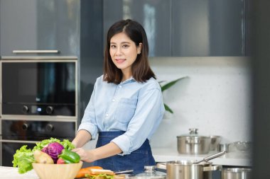 Mutfakta kameraya bakan Asyalı kadın. Sebzeler bol ekmek ve yemek masasında. Şef aşçılık konsepti..