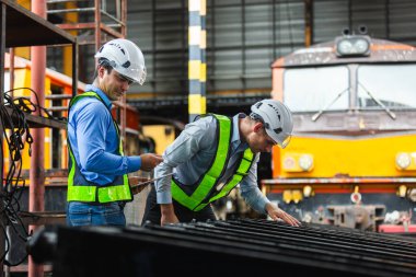Güvenlik teçhizatı takan mühendisler, kontrol listesi ve elle inceleme kullanarak tren istasyonundaki mekanik parçaları inceliyorlar. Endüstriyel bakım, teknik denetim ve kalite kontrolü kavramı.