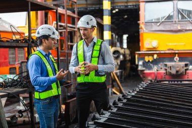 Güvenlik teçhizatı takan mühendisler, kontrol listesi ve elle inceleme kullanarak tren istasyonundaki mekanik parçaları inceliyorlar. Endüstriyel bakım, teknik denetim ve kalite kontrolü kavramı.