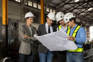 Bir grup demiryolu mühendisi ve güvenlik kaskı takmış iş adamları bir sanayi fabrikasının planlarını gözden geçiriyorlar. Proje planlama, beyin fırtınası ve mühendislik takım çalışması kavramı.
