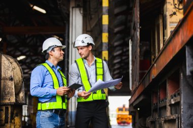 Güvenlik kaskı ve yansıtıcı yelek takan mühendisler, demiryolu ekipmanlarını taslak ve dijital tablet kullanarak teftiş ediyorlar. Endüstriyel denetim, mühendislik planlama ve takım çalışması kavramı.