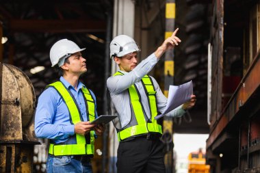 Güvenlik kaskı ve yansıtıcı yelek takan mühendisler, demiryolu ekipmanlarını taslak ve dijital tablet kullanarak teftiş ediyorlar. Endüstriyel denetim, mühendislik planlama ve takım çalışması kavramı.