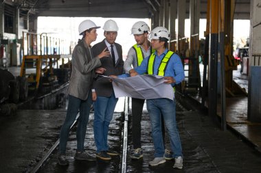Bir grup demiryolu mühendisi ve güvenlik kaskı takmış iş adamları bir sanayi fabrikasının planlarını gözden geçiriyorlar. Proje planlama, beyin fırtınası ve mühendislik takım çalışması kavramı.