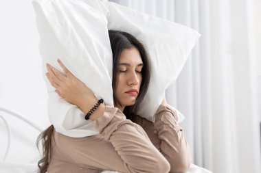 Yatakta otururken kulaklarını yastıkla kapatan genç kadınlar, hayal kırıklıklarını, uyku bozukluklarını ya da yatak odasındaki gürültü sorunlarını ifade ediyorlar, uykusuzluk ve sabah rahatsızlıklarını vurguluyorlar..