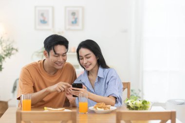 Asyalı çift evde akıllı telefon kullanırken kahvaltıdan zevk alıyor. Mutlu ilişki kavramı, dijital yaşam tarzı, iletişim ve rahat mutfakta modern sabah rutini..