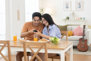 Mutlu Asyalı çift kahvaltıdan ve rahat ev ortamında akıllı telefonlardan keyif alıyor. Aşk kavramı, bağlantı, dijital yaşam tarzı ve yemek masasında rahatlatıcı sabah rutini..
