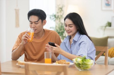 Genç Asyalı çift evde kahvaltı yapıyor. Kadınlar akıllı telefon kullanırken erkekler portakal suyu içiyor. Konforlu yaşam tarzı, dijital rutin ve evcil ilişkilerdeki anlar.