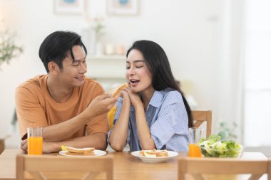 Asyalı bir adam kahvaltıda kız arkadaşına sandviç yediriyor. Aşk, ilgi ve mutluluk dolu romantik bir an. Konforlu mekanda ilişki bağı ve dijital çift yaşam tarzı kavramı.