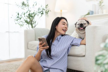 Asyalı kadınlar gülümserken akıllı telefon kullanıyor ve rahat oturma odasında evcil köpeğe sarılıyorlar. Mutlu dijital yaşam biçimi, evcil hayvan arkadaşlığı, ev içi sağlık ve rahatlama ortamı kavramı.