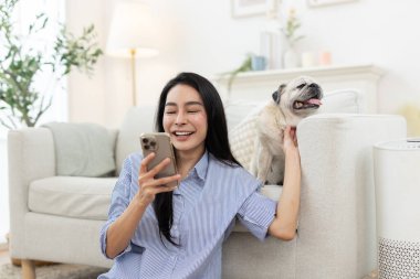 Asyalı kadınlar gülümserken akıllı telefon kullanıyor ve rahat oturma odasında evcil köpeğe sarılıyorlar. Mutlu dijital yaşam biçimi, evcil hayvan arkadaşlığı, ev içi sağlık ve rahatlama ortamı kavramı.