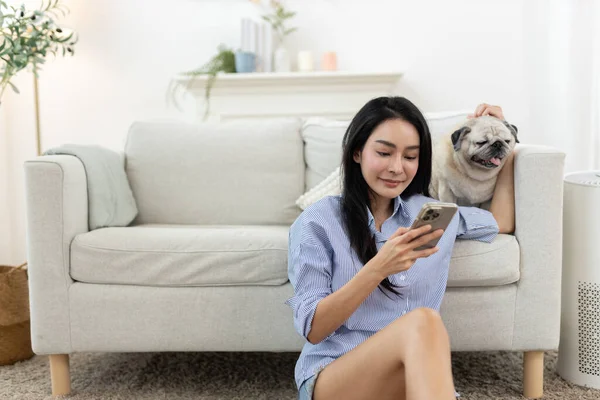 Asyalı kadınlar gülümserken akıllı telefon kullanıyor ve rahat oturma odasında evcil köpeğe sarılıyorlar. Mutlu dijital yaşam biçimi, evcil hayvan arkadaşlığı, ev içi sağlık ve rahatlama ortamı kavramı.