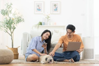 Evde dizüstü bilgisayar ve akıllı telefon kullanan Asyalı çift halının üzerinde rahatlayan evcil köpekle birlikte. Dijital yaşam tarzı kavramı, evden çalışma, teknoloji, ilişki ve rahat ev hayatı