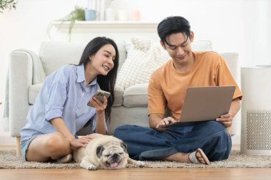 Evde dizüstü bilgisayar ve akıllı telefon kullanan Asyalı çift halının üzerinde rahatlayan evcil köpekle birlikte. Dijital yaşam tarzı kavramı, evden çalışma, teknoloji, ilişki ve rahat ev hayatı