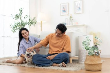 Mutlu Asyalı çift oturma odasında sevimli bir köpekle oynuyor. Aşk kavramı, ilişki bağı, aile hayatı, evcil hayvan arkadaşlığı ve modern evdeki neşeli anlar..