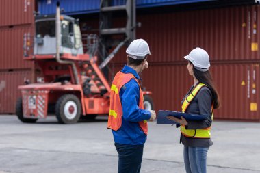 Güvenlik yeleği giyen iki lojistik uzmanı nakliye sahasında konteyner taşıyıcısının yanında yük taşımayı tartışıyor, takım çalışması, kargo operasyonları ve endüstriyel planlamayı temsil ediyorlar..