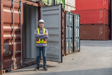 Güvenlik yeleği ve beyaz kask kollu Asyalı işçiler nakliye deposundaki açık kargo konteynerinin önünde durarak lojistik, ihracat, ithalat ve sanayi tedarik zincirini sembolize ediyor..