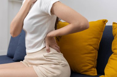 Kanepede otururken belden aşağısı ağrıyan genç kadınların yakın çekimleri, muhtemelen kötü duruş, uzun oturma saatleri veya kas gerilmesinden kaynaklanıyor. Sırt ağrısı, sağlık sorunu veya ofis sendromu kavramı.