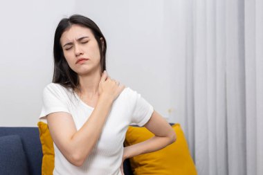 Omuz ve boyun ağrısından muzdarip genç kadınlar, muhtemelen kötü duruş veya stres yüzünden, evdeki kanepede oturuyorlar. Kas gerilimi, işle ilgili gerginlik veya yerleşik yaşam tarzı kavramı.