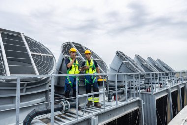 Güvenlik teçhizatında iki mühendis çatı soğutma kulesi sistemlerini kontrol ederek bakım ve enerji optimizasyonunu sağlıyor. HVAC teknolojisi, tesis yönetimi ve endüstriyel işletme kavramı.