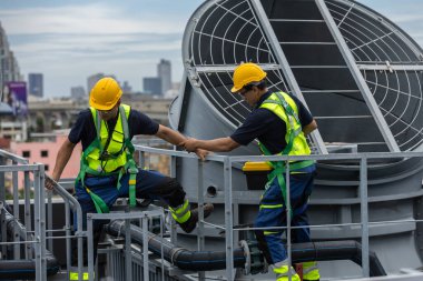 Güvenlik teçhizatı takan iki mühendis çatıdaki havalandırma borularından geçerken birbirlerine yardım ediyorlar. Tehlikeli iş ortamlarında takım çalışması, endüstriyel güvenlik, tesis bakımı ve destek kavramı.