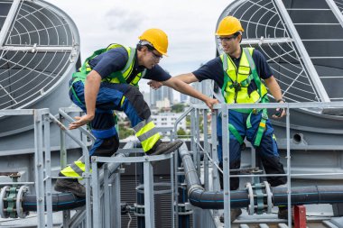 Güvenlik teçhizatı takan iki mühendis çatıdaki havalandırma borularından geçerken birbirlerine yardım ediyorlar. Tehlikeli iş ortamlarında takım çalışması, endüstriyel güvenlik, tesis bakımı ve destek kavramı.