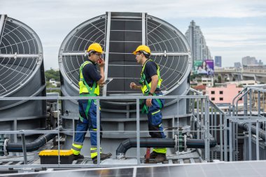 Güvenlik donanımlı iki mühendis tabletteki verileri kontrol ederken HVAC sistem operasyonunu tartışıyor. Akıllı tesis yönetimi, endüstriyel bakım ve çatıda mekanik denetim kavramı.
