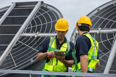 Güvenlik donanımlı iki mühendis tabletteki verileri kontrol ederken HVAC sistem operasyonunu tartışıyor. Akıllı tesis yönetimi, endüstriyel bakım ve çatıda mekanik denetim kavramı.