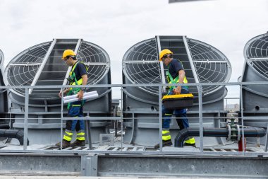 Güvenlik teçhizatı olan mühendisler çatıdaki havalandırmayı ve güneş paneli sistemlerini enerji verimliliği için inceliyorlar. Endüstriyel bakım, temiz enerji ve tesis yönetimi kavramı.