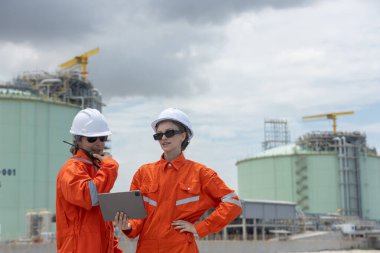 Turuncu üniformalı ve kasklı iki mühendis dijital tablet ve radyoyla bir gaz santralinde proje verilerini tartışıyor, takım çalışması, teknoloji, enerji endüstrisi ve saha operasyonlarını temsil ediyorlar..