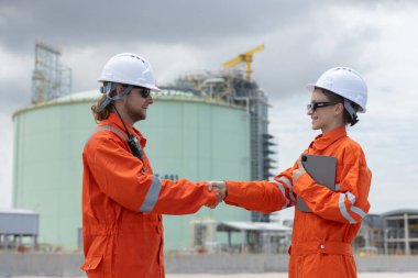 Turuncu güvenlik üniforması ve beyaz kasklı iki mühendis endüstriyel gaz fabrikasında el sıkışıyor, ortaklığı, takım çalışmasını ve mühendislik operasyonlarında başarılı işbirliğini simgeliyor..
