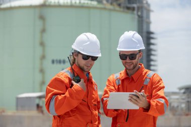 Turuncu güvenlik üniforması ve kasklı iki mühendis, bir gaz veya enerji tesisindeki saha operasyonlarını incelemek için dijital tablet kullanıyorlar, takım çalışması, iletişim ve endüstriyel denetimi gösteriyorlar..
