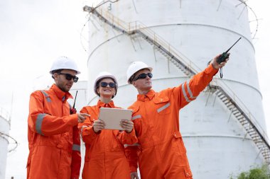 Bir petrol rafinerisinde güvenlik üniforması ve kask giyen bir grup mühendis, denetim ve operasyon görevlerini koordine etmek için tablet ve telsiz kullanıyor..