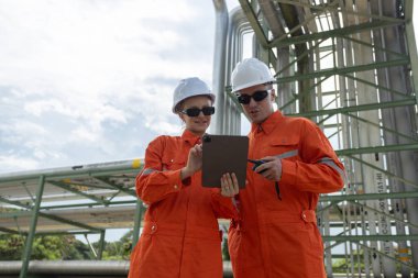 Turuncu güvenlik üniforması ve kalın şapkalı iki endüstri mühendisi dijital tablet kullanarak boru hattı sistemlerini inceliyor. Bir enerji veya petrol tesisinde saha çalışması, güvenlik ve takım çalışması.