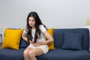 Kanepede oturan ve karın bölgesini ağrı ifadesiyle tutan kadınlar adet sancıları, karın ağrısı ya da sindirim sorunları belirtileri gösteriyorlar. Kadın sağlığı ve rahatsızlığı kavramı.