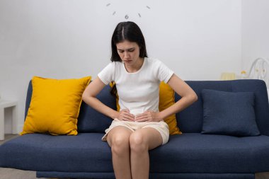 Kanepede oturan ve karın bölgesini ağrı ifadesiyle tutan kadınlar adet sancıları, karın ağrısı ya da sindirim sorunları belirtileri gösteriyorlar. Kadın sağlığı ve rahatsızlığı kavramı.