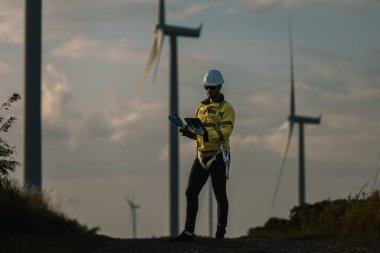 Günbatımında rüzgâr tarlasında tablet kullanan mühendis, akıllı enerji izleme, dijital denetim ve yenilenebilir enerji ve temiz teknolojide sürdürülebilir gelişime bağlılığı temsil ediyor..