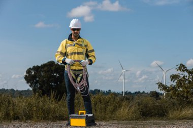 Güvenlik teçhizatı mühendisi tırmanma ipi tutuyor ve rüzgar çiftliğinde alet çantasının yanında duruyor. Yenilenebilir enerji bakımı, alan güvenliği ve temiz enerji operasyonlarını temsil ediyor..