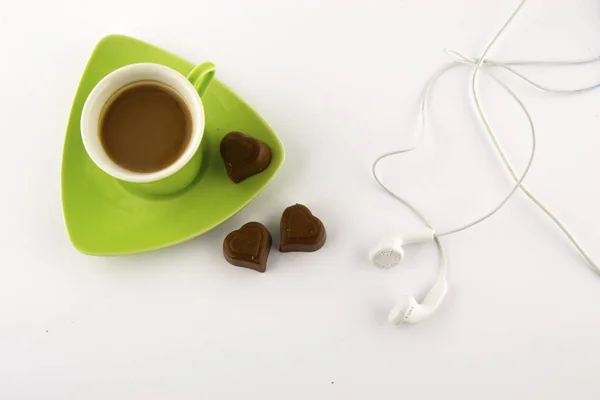 Coffee in a green cup with heart-shaped chocolate and headphones on a white background. Top view.